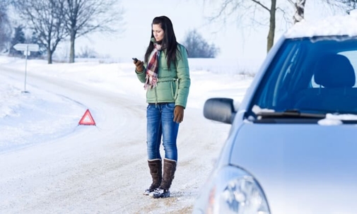A woman in winter clothing is standing next to a car on a snowy road, using a phone.